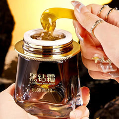 Glass jar of black diamond cream with a hand pouring cream, against a yellow background.