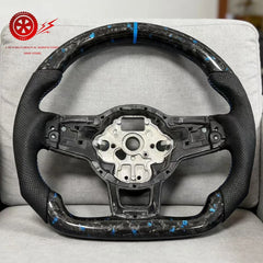 Carbon fiber steering wheel on a gray cushion with a brand logo in the corner.